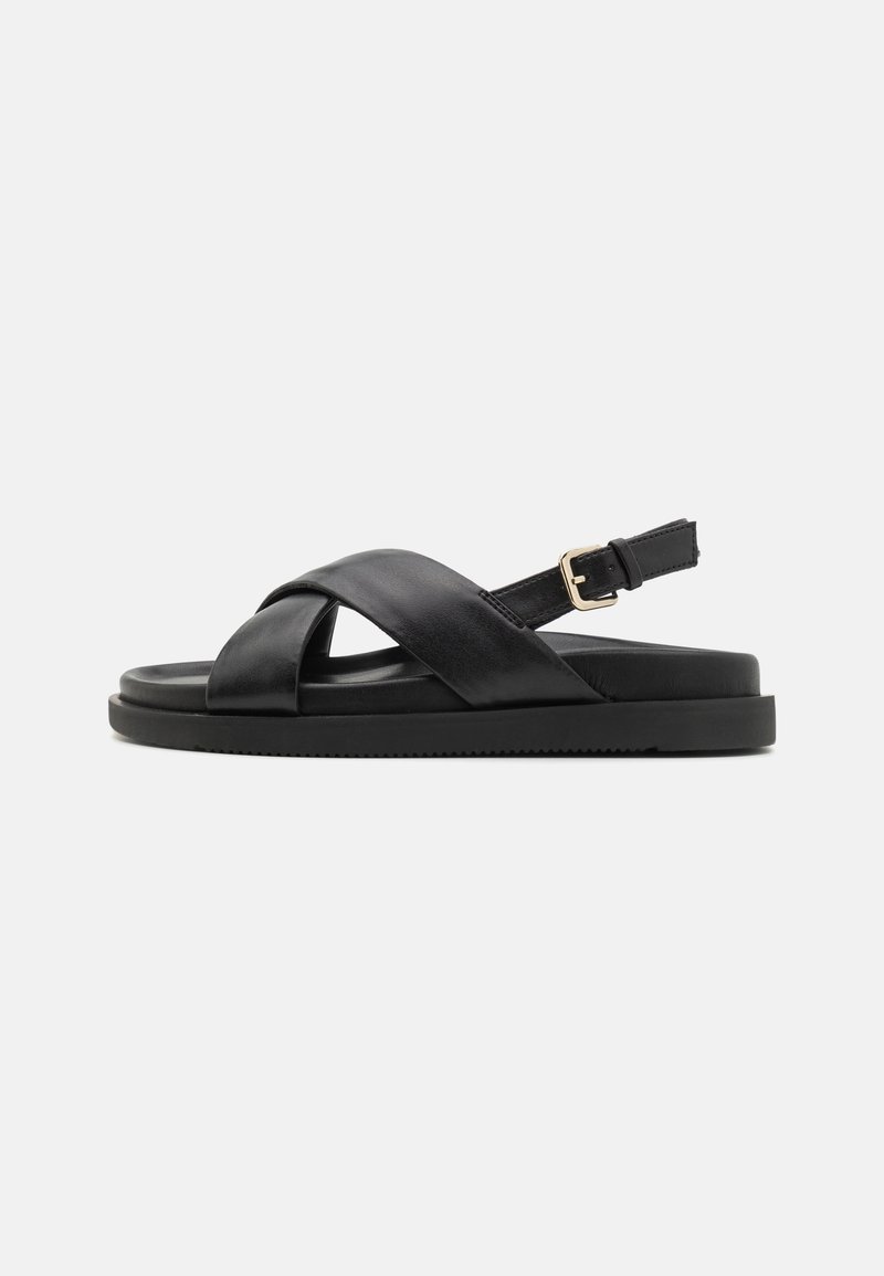 Sandale plate en cuir noir avec des brides croisées et une boucle ajustable à la cheville sur fond uni.