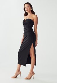 Schwarzes schulterfreies Kleid mit gerafftem Design, schrägem Schlitz bis zum Knie und eleganter Textur. Trägt dazu nudefarbene High-Heel-Sandalen.