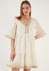 Robe crème claire avec un décolleté en V, des motifs brodés et des détails en œillets. Elle présente des manches évasées et un ourlet en couches avec volants.