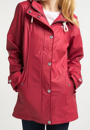 Femme portant un imperméable rouge avec des boutons à l'avant et des poches, des cordons blancs sur la capuche, et les mains dans les poches.