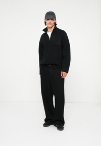 Pulover negru din fleece cu fermoar pe un sfert, buzunar pe piept, purtat cu pantaloni negri lejeri și pantofi negri, pe un fond alb simplu.