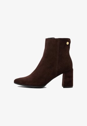 Sötétbarna velúr bokacsizma, vastag sarokkal és kis fém márkajelzéssel a felső széleinél, oldalnézet fehér háttérrel.