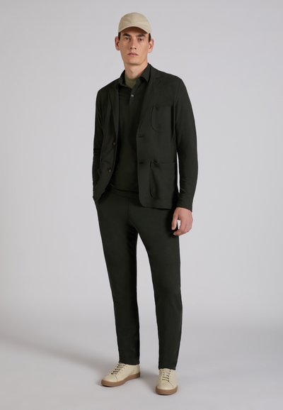 Mężczyzna w czarnej marynarce, czarnych spodniach, oliwkowej koszuli, beżowej czapce i beżowych trampkach stoi na jednolitym szarym tle.