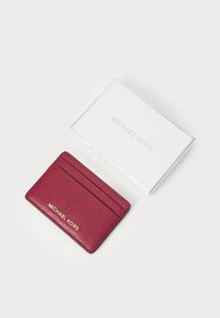 Karmosinrød skinnkortholder med teksturert overflate, med tre spor og gullpræget "Michael Kors"-logo, plassert nær en hvit boks.