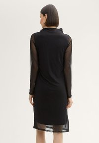 Schwarzes Kleid mit langen durchsichtigen Ärmeln, einem offenen Rückenausschnitt und einem knielangen Saum. Glattes Material, elegante Form, minimalistischer Stil.