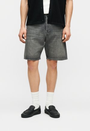 Person trägt graue Jeansshorts, weiße Socken, schwarze Slipper, schwarze Knopfweste und weißes Hemd, steht vor einem schlichten Hintergrund.
