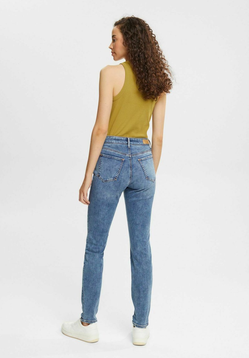 Eng anliegende blaue Jeans mit einer hohen Taille und Gesäßtaschen, kombiniert mit einem senfgelben Tanktop und weißen Sneakers. Lockige Haare sind zu sehen.