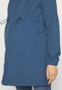 Gros plan d'une veste zippée bleue avec poignets élastiques, cordons ajustables à la taille et poches latérales portée sur un jean.