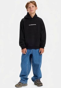 Μαύρο φούτερ με κουκούλα, τσέπη μπροστά και κορδόνια στην κουκούλα; συνδυασμένο με φαρδιά μπλε τζιν και καφέ αθλητικά παπούτσια; απλό φόντο.