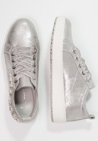 Para srebrzystych, błyszczących tenisówek damskich z satynowymi sznurówkami i perłowymi koralikami, na białych gumowych podeszwach, pokazanych z widoku z góry i z boku.