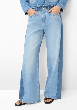 Lyseblå jeans med vid ben og mørkere sidepaneler, båret med hænderne i lommerne og sorte sandaler med åbne tæer og guldfarvet spænde.