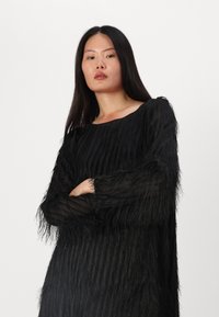 Abito nero con dettagli di frange testurizzate, maniche lunghe e scollatura ampia. Realizzato in materiale trasparente con una silhouette fluida.