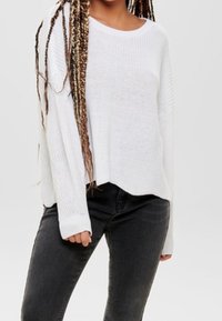 Pull en maille côtelée blanche, avec un large col et des épaules tombantes, associé à un jean noir ajusté. Tissu texturé et coupe décontractée.