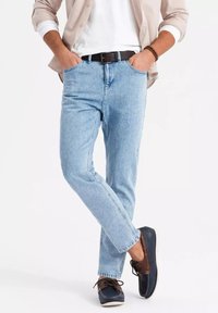 Jeans de mezclilla azul claro con diseño de pierna recta, cinco bolsillos, ligero desgaste, usados con una camisa blanca y una camisa de botones beige, complementados por zapatos oscuros.