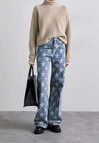 Person som bär en beige ribbad tröja, högmidjade blå jeans med vita stjärnmönster, svarta skor och håller en svart handväska.