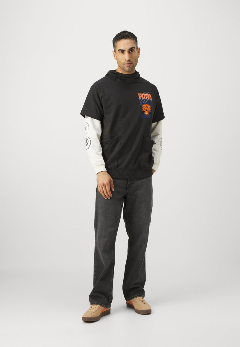 Sudadera con capucha negra y gráficos en naranja y azul, usada sobre una camiseta blanca de manga larga; combinada con jeans oscuros y zapatillas beige.