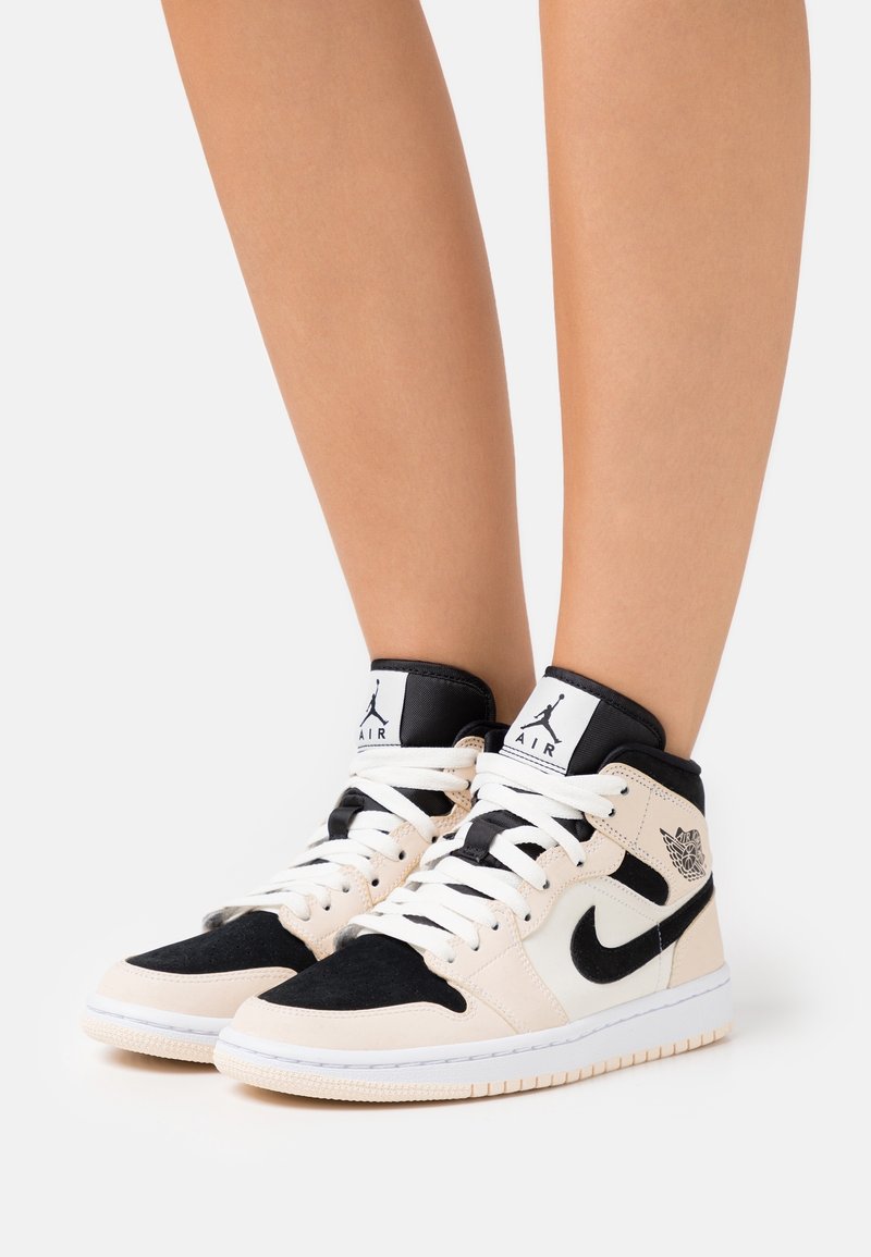 Jambes portant des baskets Air Jordan 1 mid-top beiges, noires et blanches avec des lacets blancs sur un fond clair uni.