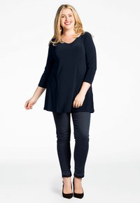 Marineblauwe tuniek met V-hals en driekwart mouwen, gecombineerd met donkere skinny jeans en zwarte hoge hakken. Glad stof, relaxte pasvorm.