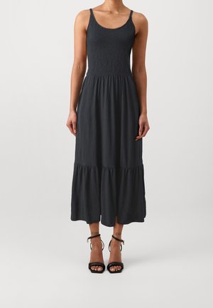 Frau trägt ein ärmelloses schwarzes Midikleid mit gerafftem Oberteil und mehrstufigem Rock, kombiniert mit schwarzen High-Heel-Sandalen mit Knöchelriemen.