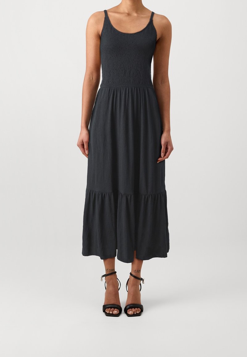 Femme portant une robe midi noire sans manches avec un corsage smocké et une jupe à volants, associée à des sandales noires à talons hauts avec bride à la cheville.