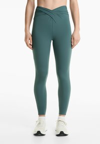 Höga, mörkgröna leggings med ett vridet mönster vid midjan, tillverkade av ett mjukt och stretchigt material. Bärs med vita träningsskor.