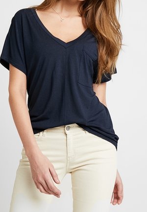 Femme portant un haut bleu marine à manches courtes avec col en V et poche poitrine, ainsi qu'un pantalon couleur crème sur un fond uni.