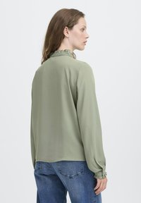 Woman facing sideways wearing a light green long-sleeve blouse with ruffled collar and cuffs paired with blue denim jeans.