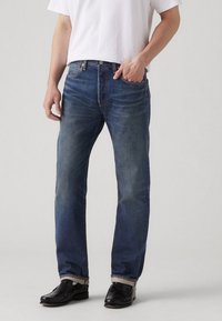 Jeans de mezclilla azul oscuro con corte recto, que presentan un diseño clásico de cinco bolsillos y costuras visibles. Llevados con zapatos negros.