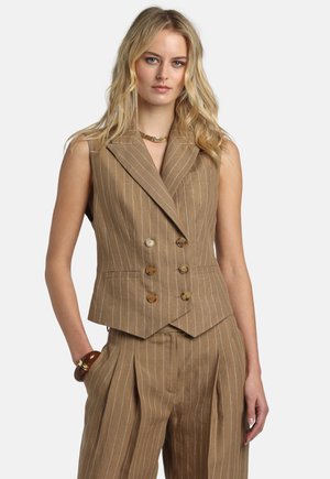 Blond kvinde iført en brun, ærmeløs, dobbeltradet vest med striber og matchende plisserede bukser, accessoriseret med en guldkæde og armbånd.