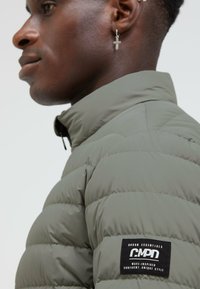Puff jacket in olive green, featuring a high collar and quilted texture. Includes a black brand patch on the left side.