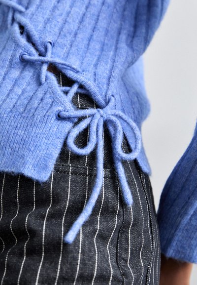 Pulover albastru cu dungi, cu detalii laterale cu șnur și fundițe, asortat cu pantaloni negri cu dungi. Accent pe textură și culoare contrastantă.