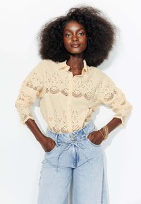 Chemise beige à boutons avec des détails en dentelle, présentant des motifs perforés. Associée à un pantalon en denim bleu clair taille haute.