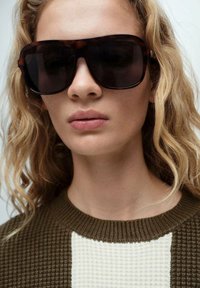 Ochelari de soare cu un cadru mare și întunecat, cu un model de broască țestoasă. Modelul poartă un pulover texturat, maro și alb. Piele netedă și păr natural.