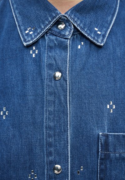 Chemise en denim bleu avec boutons-pression argentés et petits clous rectangulaires argentés disposés en motifs croisés.