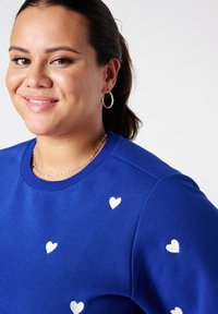 Sweat-shirt bleu avec motifs de cœurs blancs, col côtelé et texture douce. Le modèle porte des colliers en argent superposés et des boucles d'oreilles créoles.