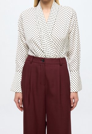 Femme portant un chemisier blanc à manches longues avec des pois noirs, rentrée dans un pantalon bordeaux taille haute à jambes larges, sur fond uni.