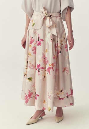 Kobieta w beżowej, kwiecistej spódnicy midi z dużą kokardą w talii, do tego beżowe czółenka na obcasie z paskiem z tyłu i noskiem w szpic.