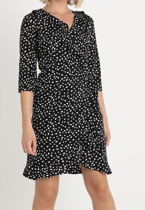 Frau trägt ein schwarzes Wickelkleid mit weißen unregelmäßigen Polka Dots, dreiviertellangen Ärmeln sowie einem Rüschenkragen und Saum.