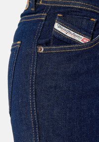 Jeans de mezclilla azul oscuro con remaches de cobre, costuras amarillas prominentes y una etiqueta visible en el bolsillo frontal. Diseño clásico de cinco bolsillos.