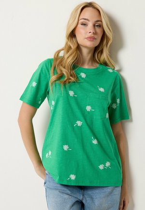 Mujer rubia con una camiseta verde de manga corta con estampados florales blancos y vaqueros azules, de pie con una mano en el bolsillo.