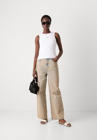 Canotta bianca con logo, jeans dritti in denim azzurro chiaro, borsa nera e infradito beige. Texture morbide e colori contrastanti.
