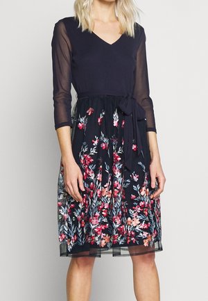 Vrouw met een zwarte jurk met doorschijnende driekwartsmouwen en een rok met bloemrijke borduursels, gestrikt in de taille met een stoffen riem.