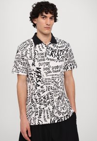 Camisa polo em preto e branco com vários logótipos e textos manuscritos. Mangas curtas, colarinho preto e um corte descontraído.