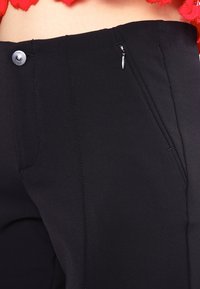 Pantalones negros hechos de una tela suave, con bolsillos frontales, cremallera y cierre de botón. Se pueden ver detalles de costura.