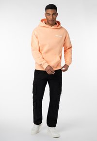 Pfirsichfarbener Hoodie mit Fronttasche und dem Text "WORLDWIDE", kombiniert mit schwarzen Cargo-Hosen mit Seitentaschen und weißen Sneakern.