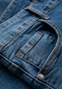 Tissu en denim bleu moyen, présentant une grande poche avec une petite poche superposée et des surpiqûres dorées contrastées, bordée par un bouton en métal.