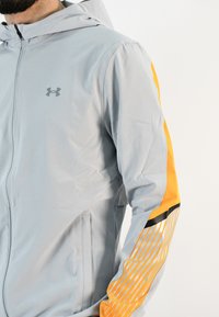 Chaqueta ligera gris con capucha, con rayas naranjas y un acento negro en la manga. Incluye cierre frontal con cremallera y bolsillos laterales.