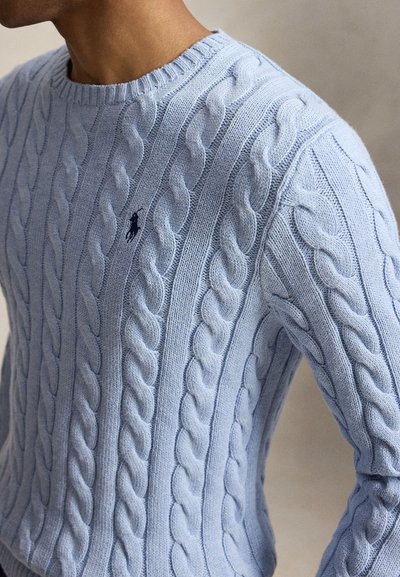 Άνδρας που φορεί ανοιχτό μπλε πουλόβερ με πλέξη câble, στρογγυλή λαιμόκοψη και μικρό σκούρο κεντημένο λογότυπο στο αριστερό μέρος του στήθους.