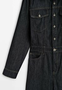 Czarny dżinsowy kombinezon z długimi rękawami, zapięcie na guziki z przodu, dwie kieszenie na klatce piersiowej z klapkami oraz kieszenie boczne z kontrastowym przeszyciem.
