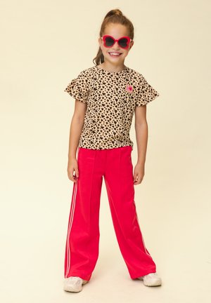 Jong meisje met rode zonnebril in de vorm van vlinders, een beige top met zwarte hartjes en felrode broek met witte strepen, glimlachend en staand.